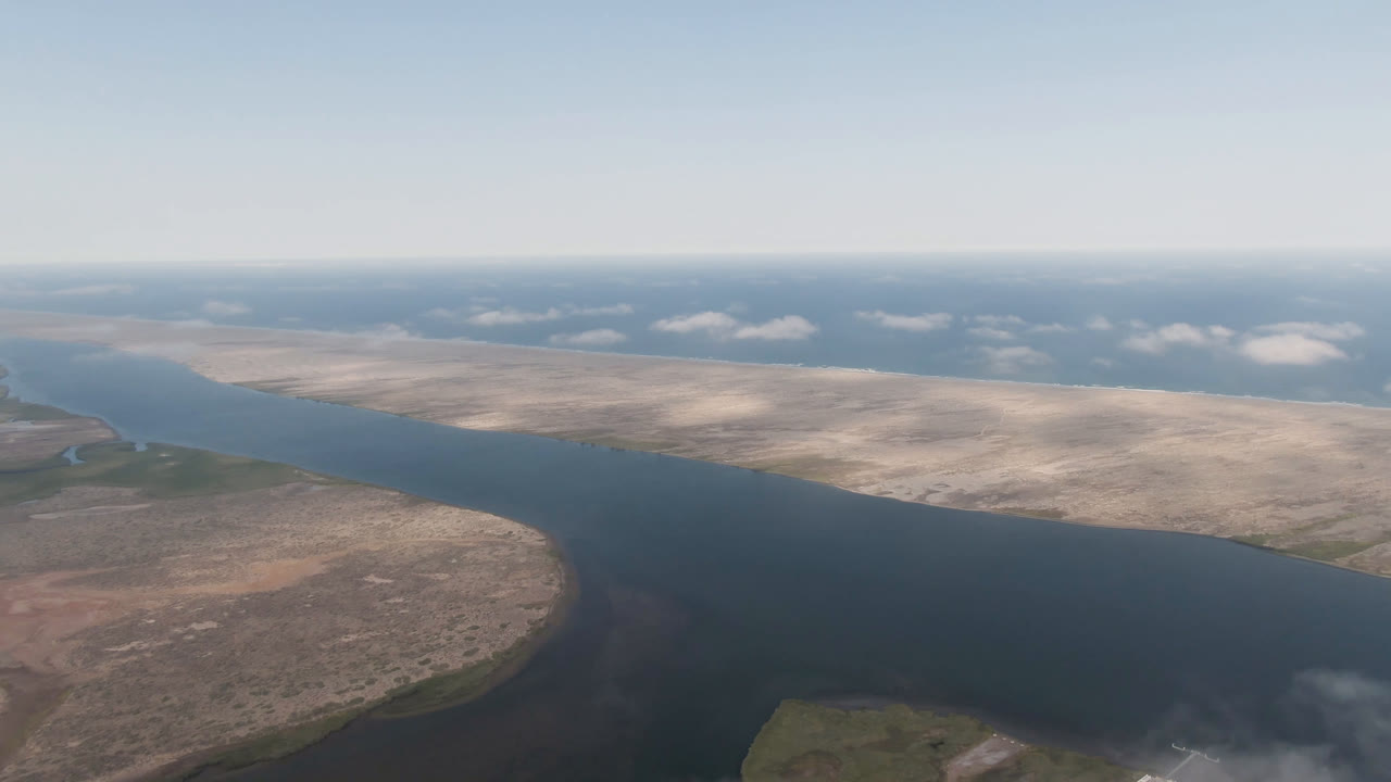 Opening aerial frame over Aldofo Lopez Mateos, Baja California Sur, with a broad dark river between tan desert flats, distant water, scattered clouds, and no visible people.