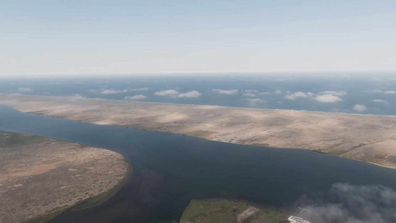 High aerial view of a long waterway with dry land on both sides and clouds in the distance.