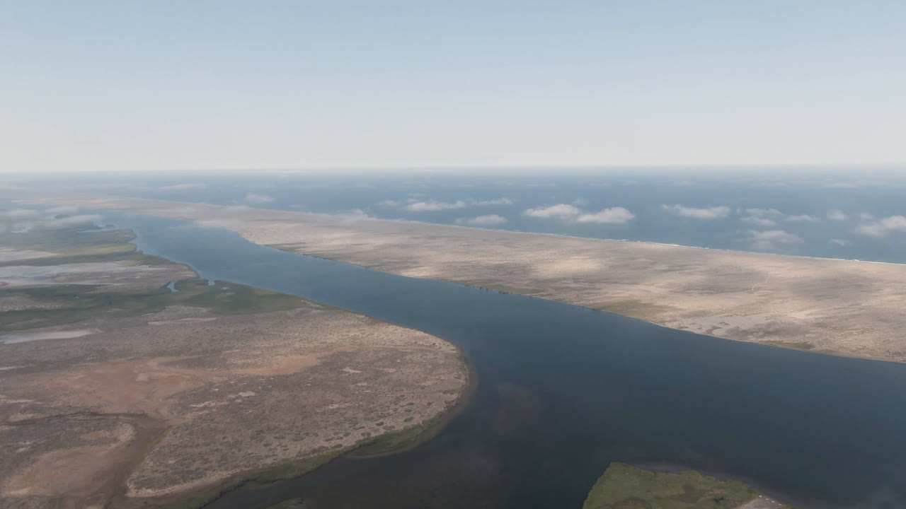 Opening aerial frame over Aldofo Lopez Mateos, Baja California Sur, with a dark river bending between tan desert banks, hazy horizon, scattered clouds, and no visible people.