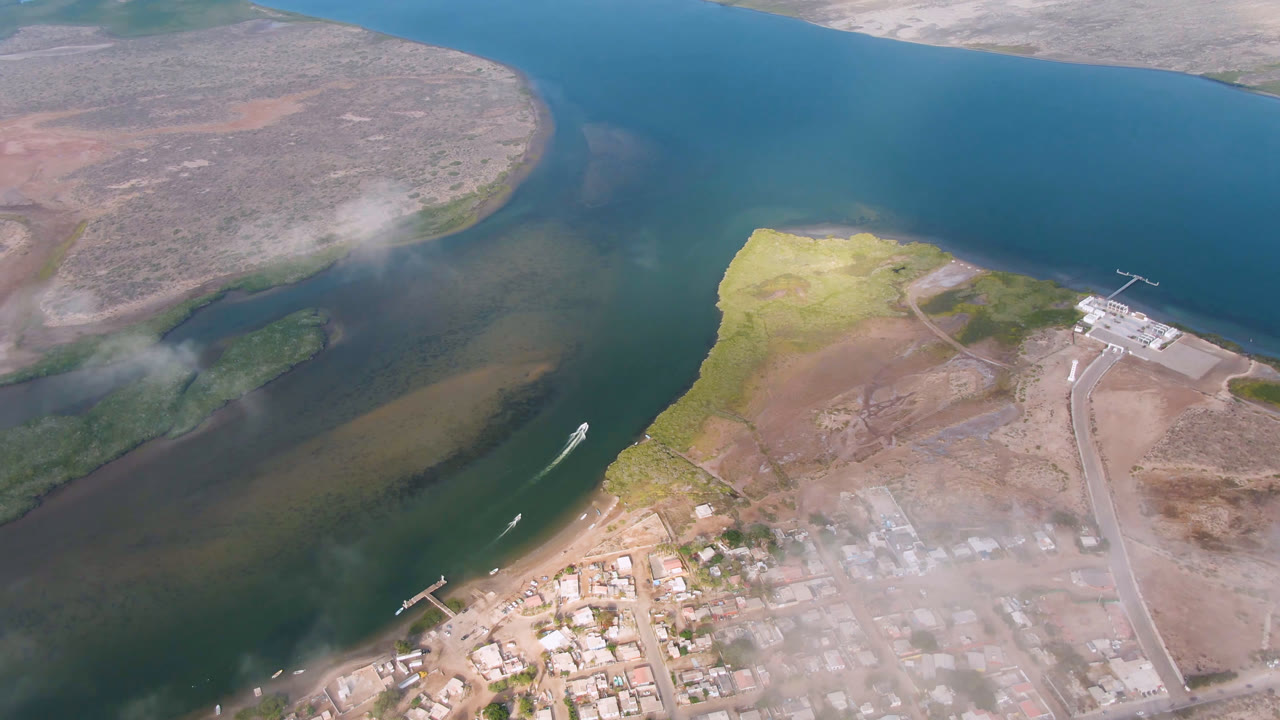 Aerial view in Aldofo Lopez Mateos with a boat wake crossing the water, docks and town buildings near the lower edge, dry land beyond, and no visible people.