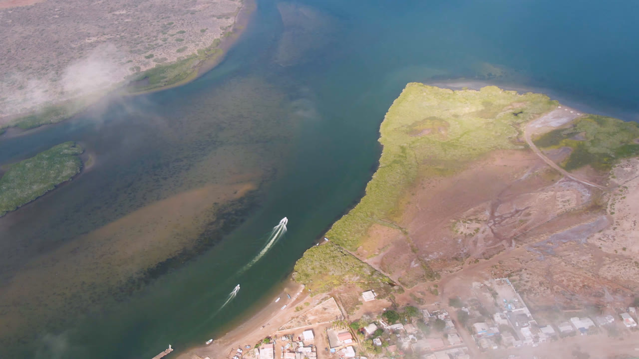 Aerial frame over Aldofo Lopez Mateos, Baja California Sur, showing a small boat leaving a white wake near sandbars, scrubland, roads, scattered buildings, and no visible people.