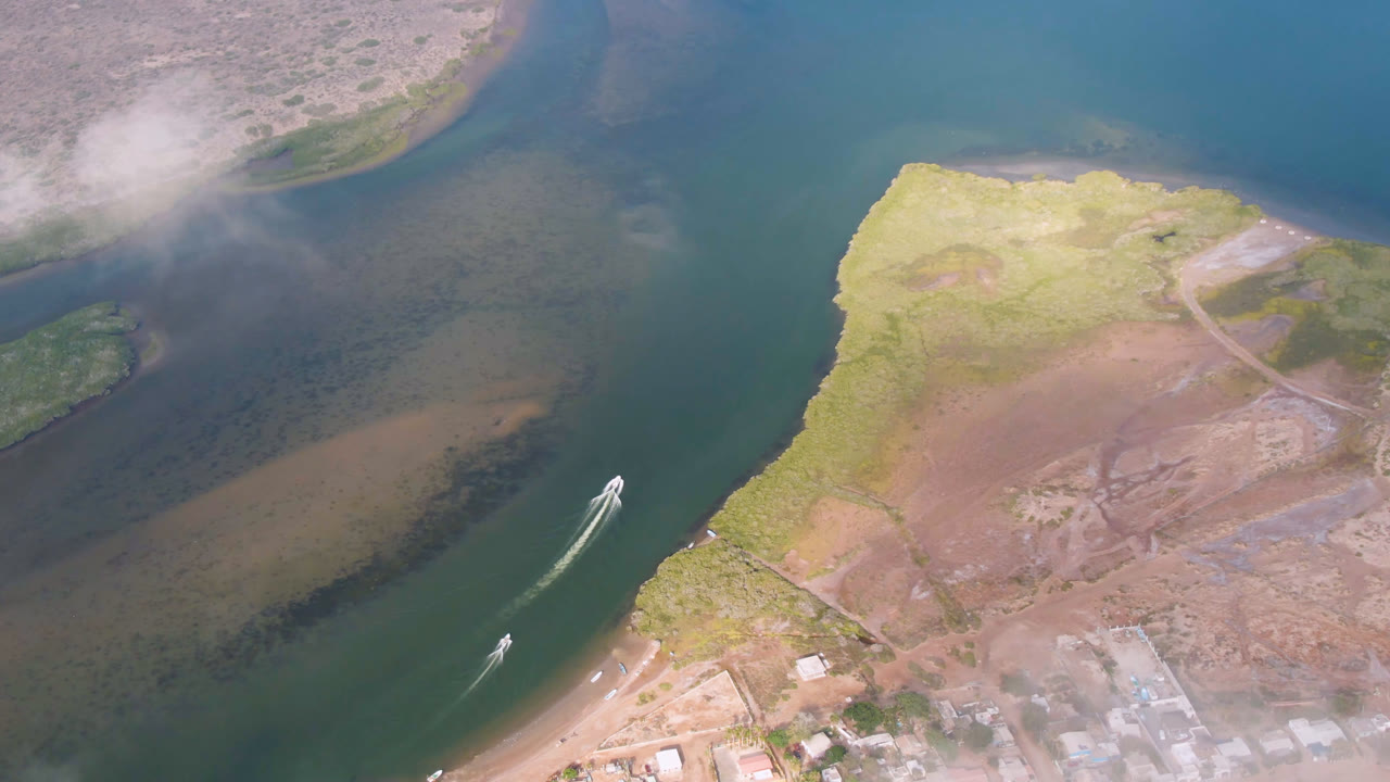 Aerial frame over Aldofo Lopez Mateos with the boat farther along the channel, a curved wake behind it, pale sand flats below, low vegetation at right, and no visible people.