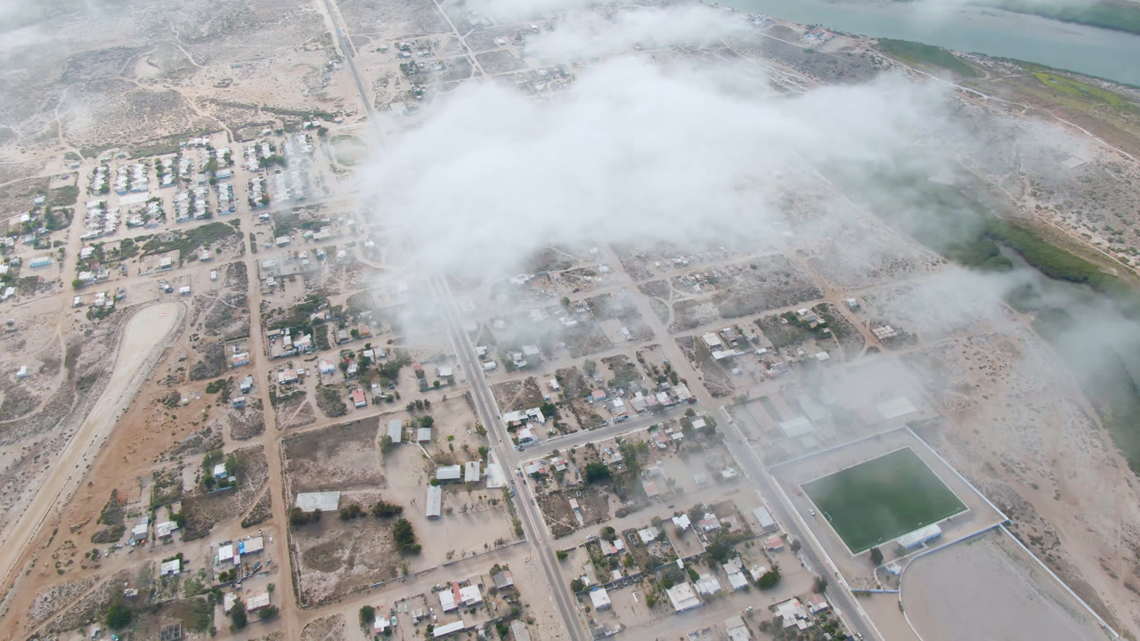 Aldofo Lopez Mateos appears from above with clustered homes, straight roads, pale shoreline water, scrubland, and a broad cloud band across the center with no visible people.