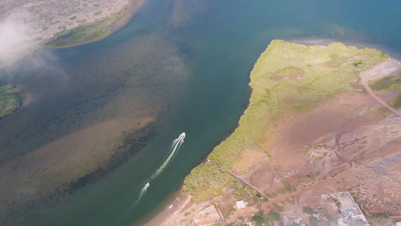 Aerial frame from Aldofo Lopez Mateos, Baja California Sur, showing a small boat beginning across green river water near pale sandbars, scrubby shoreline, and no visible people.