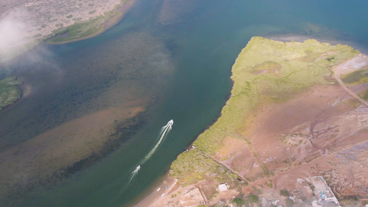 Aerial frame from Aldofo Lopez Mateos, Baja California Sur, with the boat farther along the channel, a bright wake trailing behind it, exposed shoreline to the right, and no visible people.