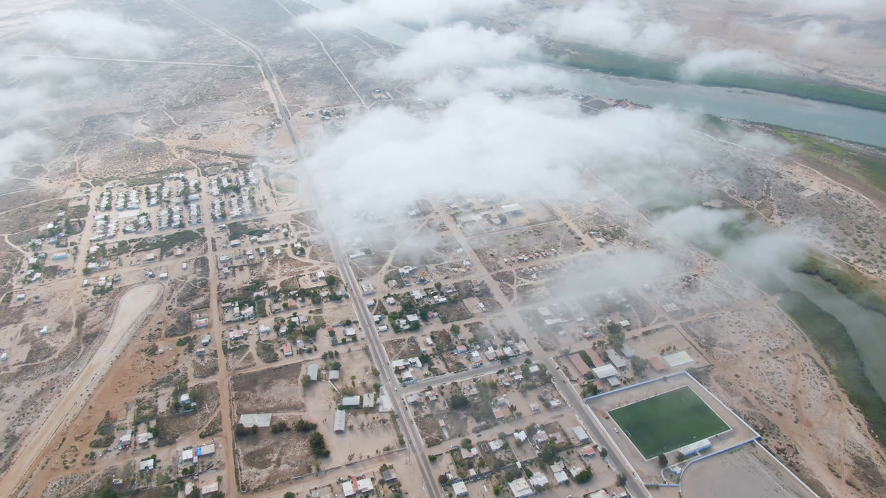 Aldofo Lopez Mateos aerial view with a straight road through town, pale desert ground, river water in the distance, and low fog crossing the center, no visible people.