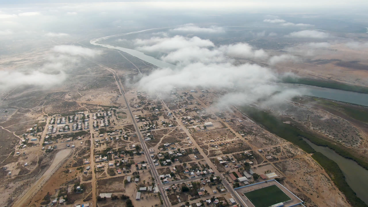 Aerial frame from Aldofo Lopez Mateos showing a straight main road through small blocks, the river curving along the right side, low cloud bands, dry terrain, and no visible people.
