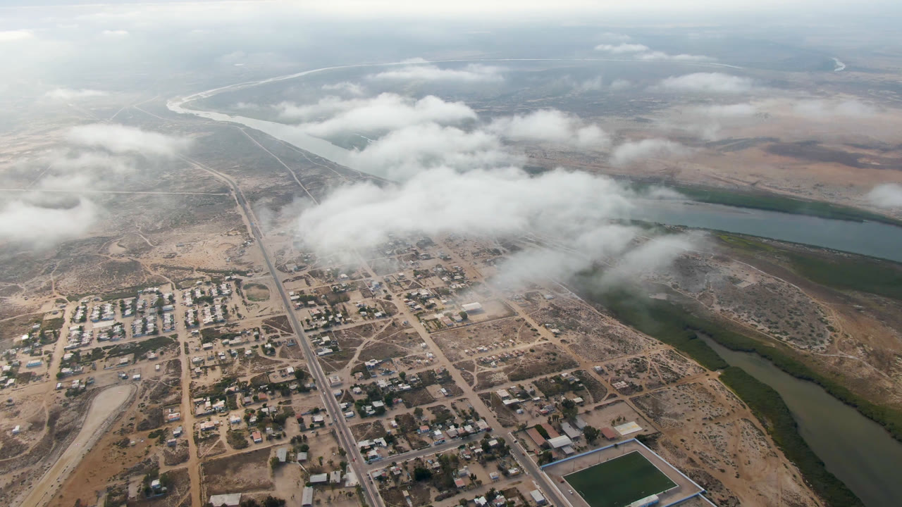 Aerial frame over Aldofo Lopez Mateos showing clustered buildings, straight roads, dry terrain, a broad river bend, low cloud cover, and no visible people.