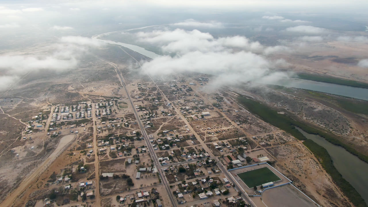 Aerial view over Aldofo Lopez Mateos, Baja California Sur, with town blocks in the foreground, a main road through the center, river water at right, low clouds overhead, and no visible people.