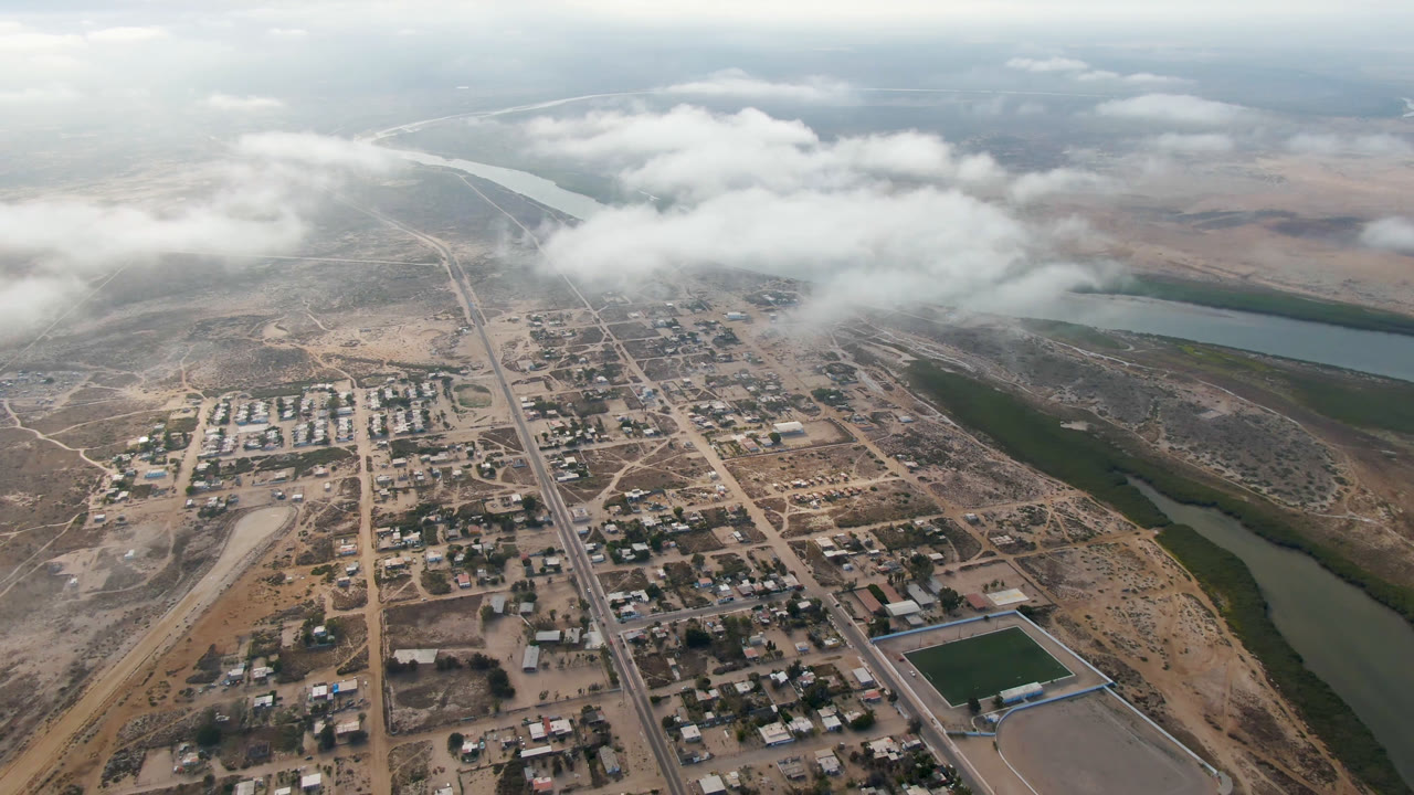 Wide aerial over Aldofo Lopez Mateos with clustered buildings and roads below, pale desert around the town, green river water to the right, cloud cover near the horizon, and no visible people.
