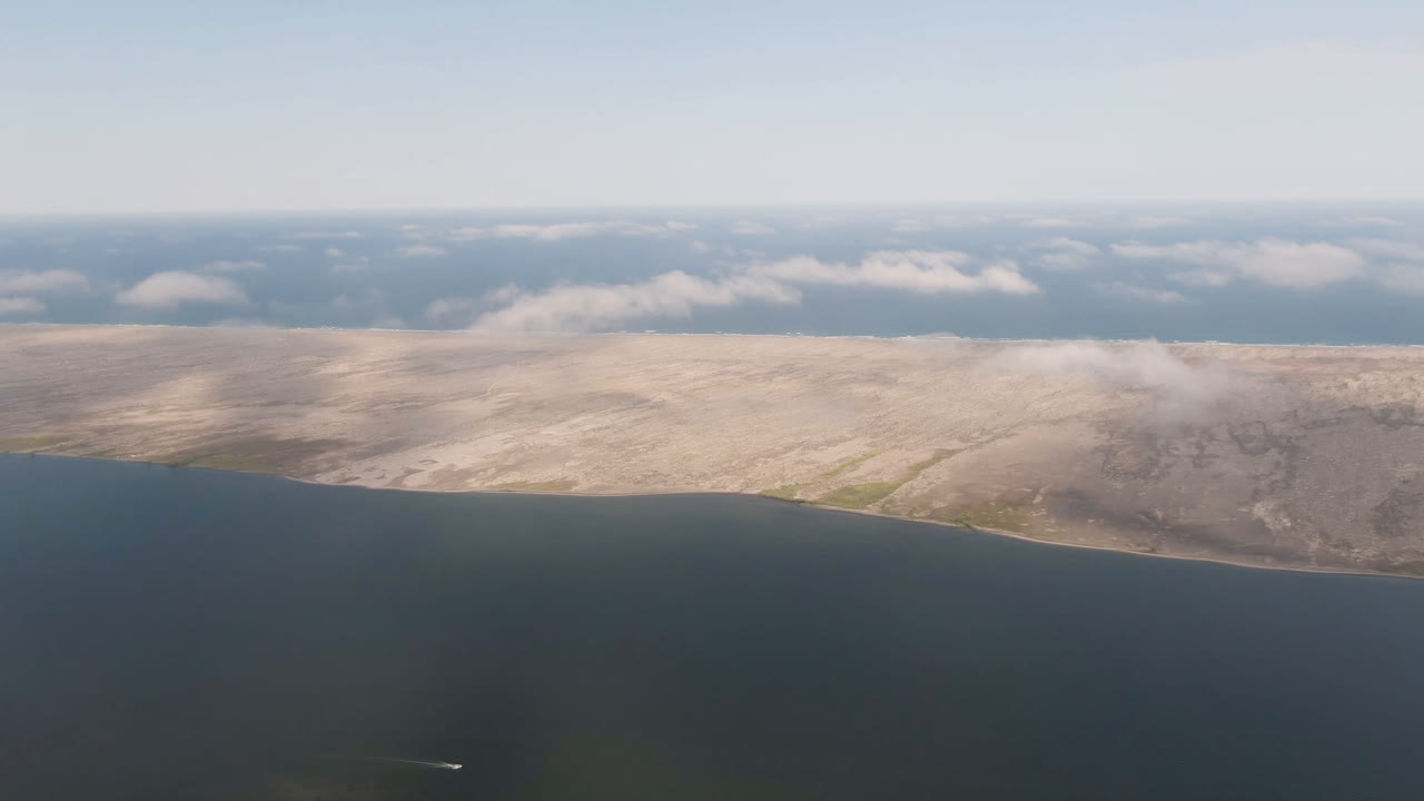 Aerial frame over dark blue water toward pale low island terrain near Aldofo Lopez Mateos, Baja California Sur, Mexico, with scattered clouds and no visible people.
