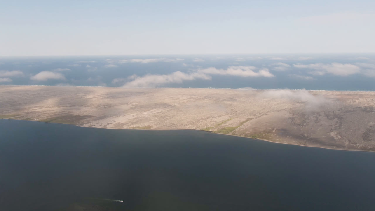 High aerial view near Aldofo Lopez Mateos, Baja California Sur, Mexico, showing dark foreground water, a pale shoreline ridge, soft clouds, and no visible people.