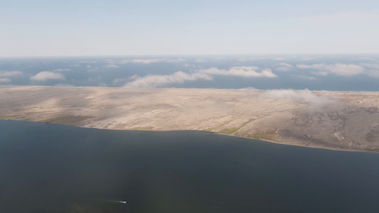 Aerial coastal frame with dark water below, dry land across the middle, and clouds beyond.