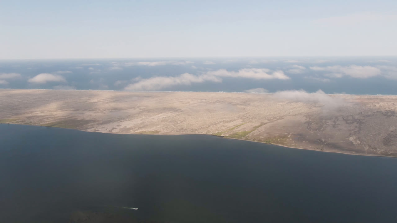 Aerial frame over dark water facing the pale Aldofo Lopez Mateos coastline in Baja California Sur, with low clouds near the horizon and no visible people.