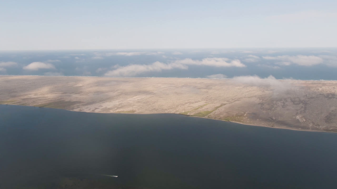 High aerial view near Aldofo Lopez Mateos, Mexico, showing dark foreground water, pale sandy land across the center, scattered cloud cover, and no visible people.