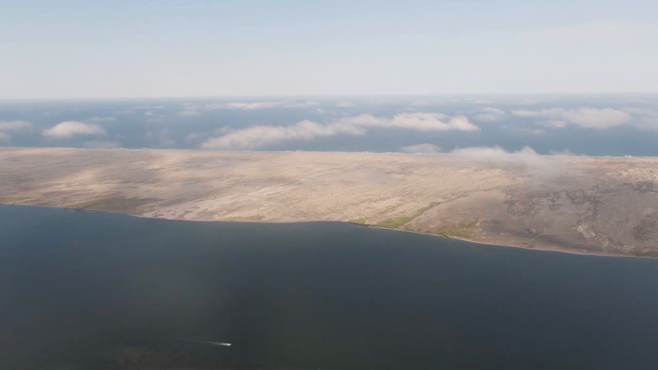 Wide aerial seascape with a long pale shoreline and cloud shadows.