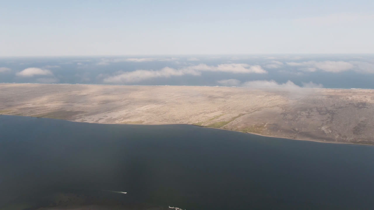 Aerial frame from Aldofo Lopez Mateos, Baja California Sur, showing dark water below, pale shoreline across the middle, low clouds on the horizon, a small white plane near the lower left, and no visible people.