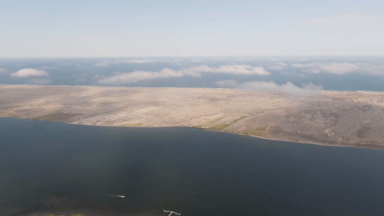 Aerial frame from Aldofo Lopez Mateos, Baja California Sur, showing dark open water, a long sandy shore, muted scrubland, a low cloud line, a small white plane near the lower center, and no visible people.