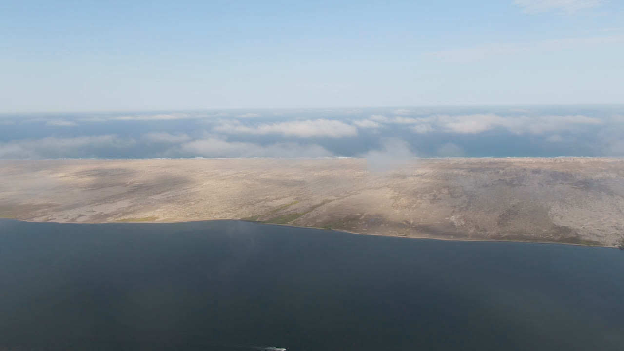 Wide aerial view near Aldofo Lopez Mateos, Baja California Sur, with dark water in the foreground, pale desert hills across the center, low clouds on the horizon, and no visible people.