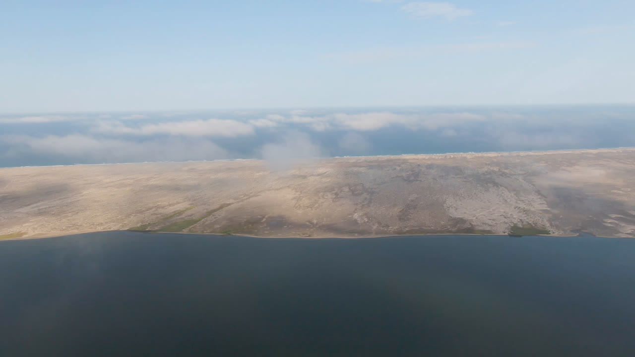 Aerial frame from Aldofo Lopez Mateos, Baja California Sur, showing dark water in the foreground, pale sandy land across the middle, low clouds on the horizon, and no visible people.