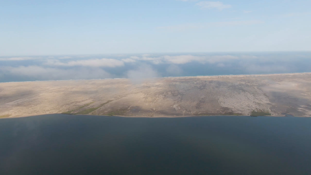 Aldofo Lopez Mateos aerial still with open dark water below, tan shoreline stretching across frame, a soft cloud band beyond the land, and no visible people.