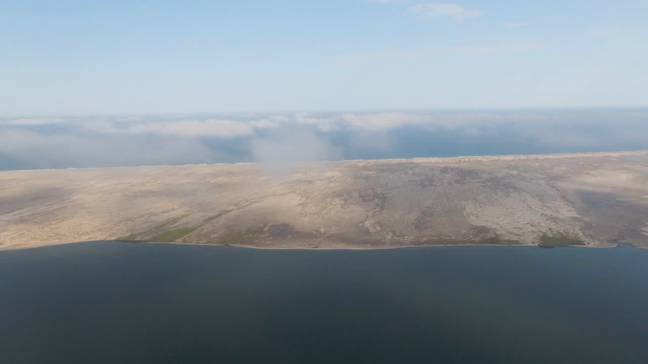 Opening aerial frame over Aldofo Lopez Mateos, Baja California Sur, with dark open water below, a pale sandy island or coastline across the center, hazy cloud along the horizon, and no visible people.