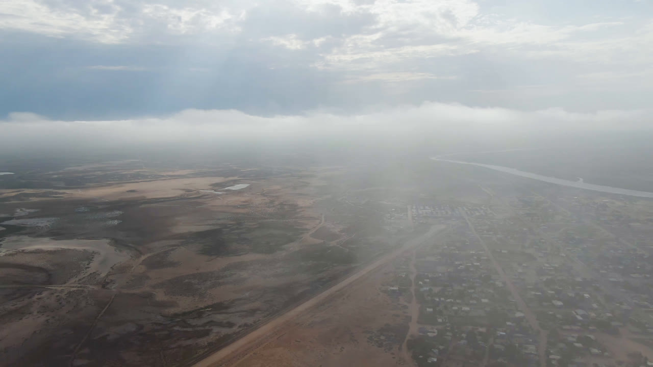 Hazy aerial view over Aldofo Lopez Mateos in Baja California Sur, with a small road grid below, dry terrain beside it, a waterway near the horizon, low clouds, and no visible people.