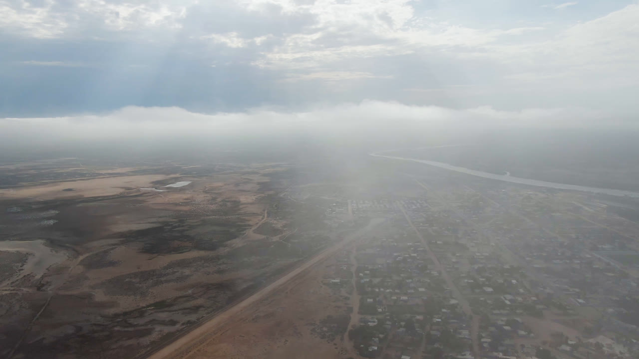 Aerial frame over Aldofo Lopez Mateos shows scattered buildings and roads softened by haze, with brown open land on both sides, a river or waterway in the distance, and no visible people.
