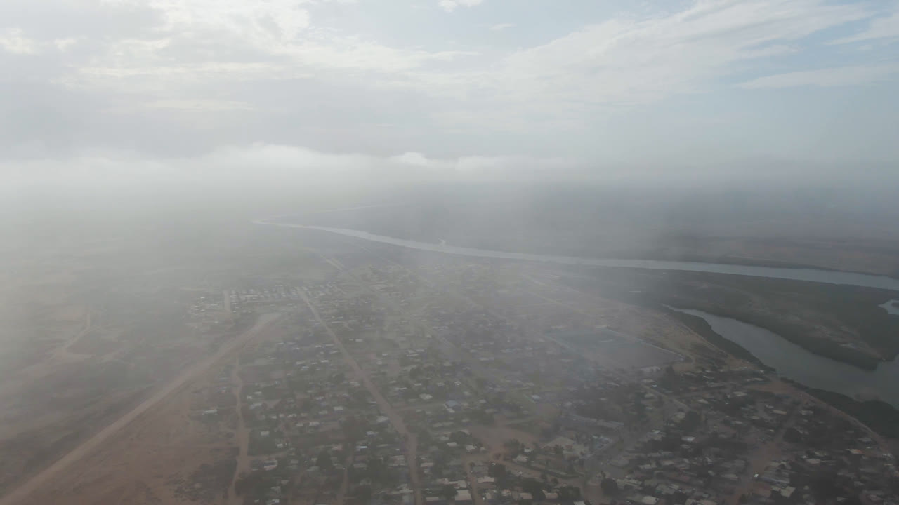 Hazy aerial view over Aldofo Lopez Mateos, Baja California Sur, with a town grid at the bottom, a river channel in the midground, cloudy sky above, and no visible people.