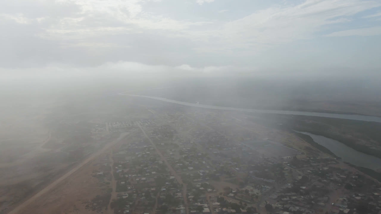 A second hazy aerial frame of Aldofo Lopez Mateos shows rooftops and pale roads below, a muted river channel beyond the town, thick fog across the center, and no visible people.