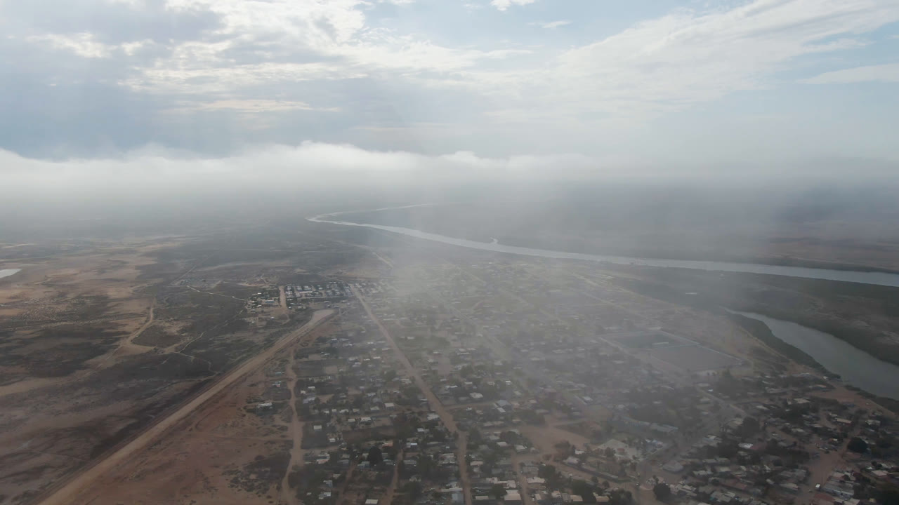 Opening aerial frame over Aldofo Lopez Mateos, Baja California Sur, showing a hazy town grid, dirt roads, river water in the distance, dry terrain, cloud cover, and no visible people.