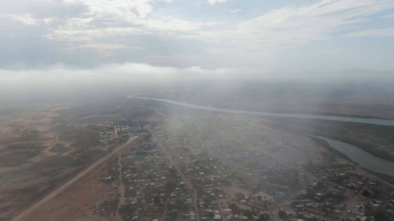 Second aerial frame in Aldofo Lopez Mateos shows the settlement below through thicker haze, with a pale river channel crossing the background and no visible people.