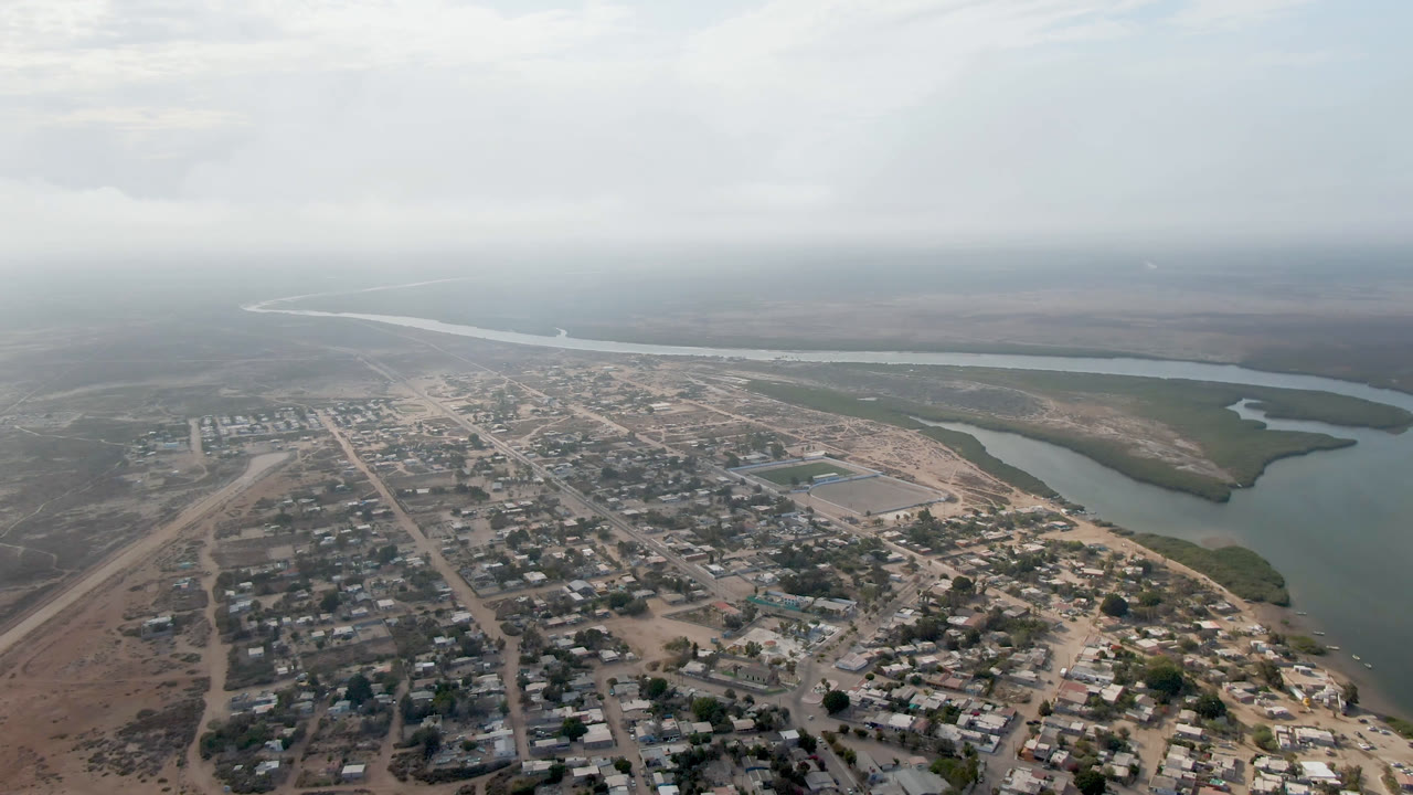 Second aerial frame in Comondu, Mexico, with the village grid centered lower in frame, a rectangular open lot near the middle, river water to the right, and no visible people.