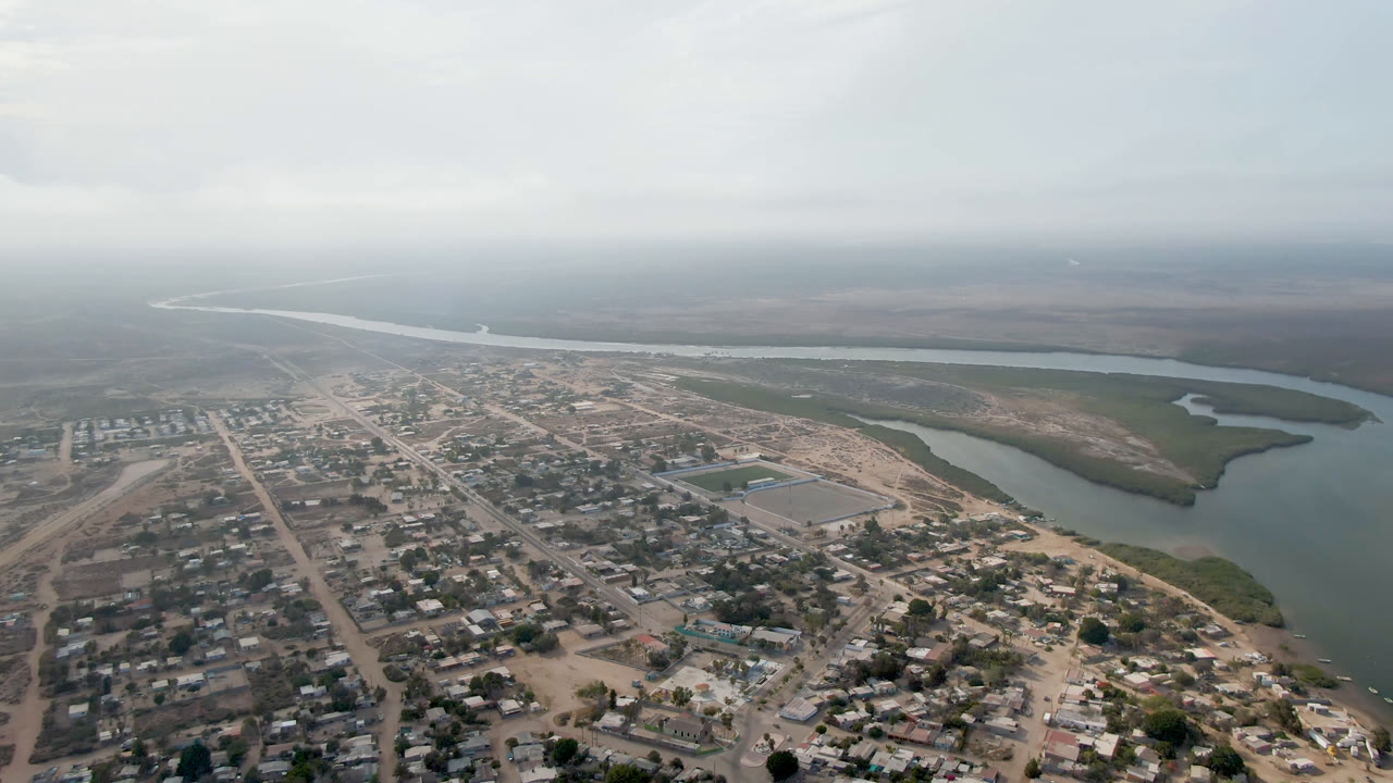 Aerial frame over Adolfo Lopez Mateos, Mexico with the dirt road grid and small structures more centered, a river bend to the right, and no visible people.