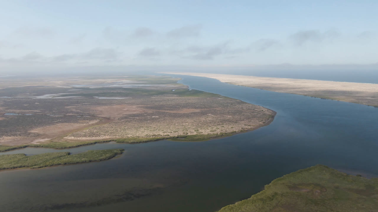 Opening aerial view over Aldofo Lopez Mateos, Baja California Sur, with a broad blue-green channel, low scrubland, pale sand flats, soft clouds, and no visible people.