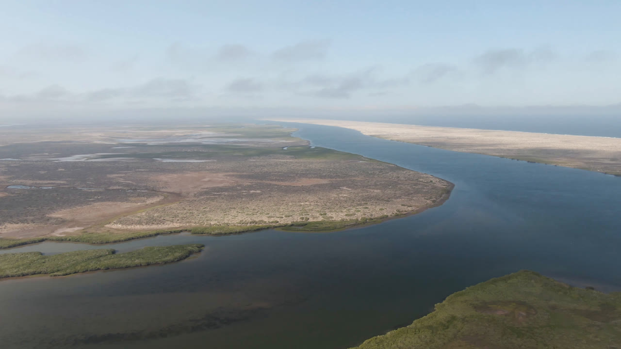 Aerial frame over Aldofo Lopez Mateos showing the channel curving past brown scrubland, shallow lagoon edges, pale sandbars, distant ocean line, and no visible people.