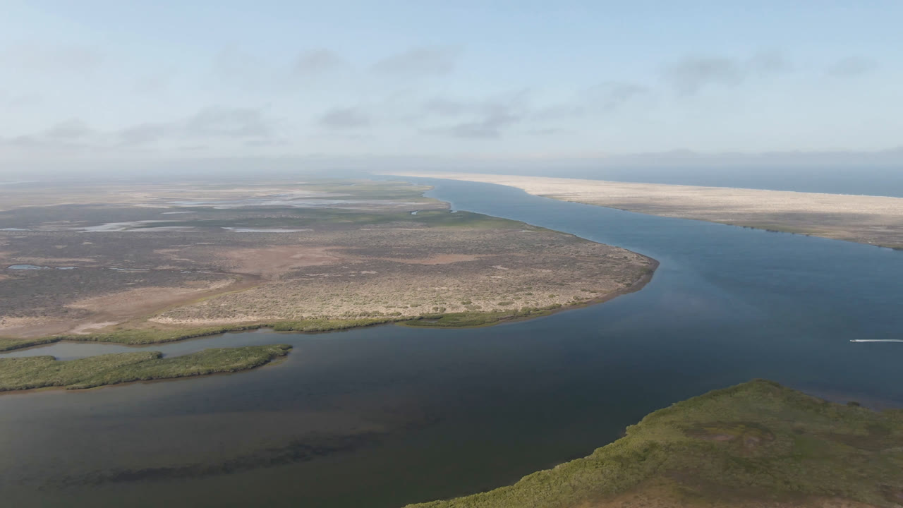 Aerial frame from Aldofo Lopez Mateos, Baja California Sur, showing broad blue water between low scrubland and pale sandy shoreline under scattered cloud, with no visible people.