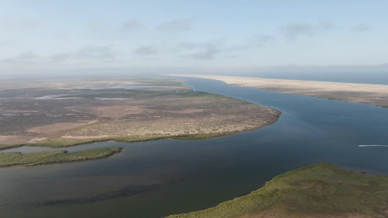 Aerial view over Aldofo Lopez Mateos water and low coastal terrain, with dark blue midground water, pale sandbars in the distance, and no visible people.