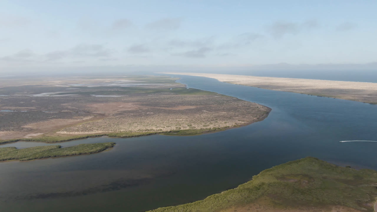 Aerial landscape with water channel, scrub foreground, sandy far shore, and a small white trace at right.