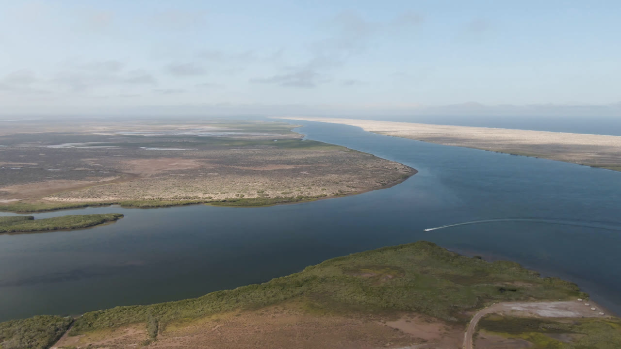 Wide aerial view of a river channel, low scrub shoreline, and distant pale coast