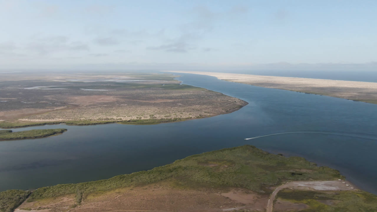 Aerial view over Aldofo Lopez Mateos, Baja California Sur, Mexico, showing the river curving past low green-brown shoreline while a small white wake appears on the water, with no visible people.