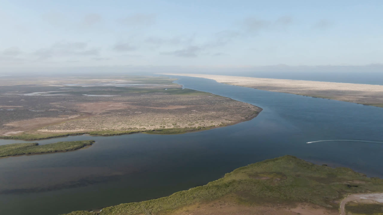 Opening aerial frame over Aldofo Lopez Mateos, Baja California Sur, with dark blue-green water between scrubland, a pale beach strip, distant ocean, scattered clouds, and no visible people.