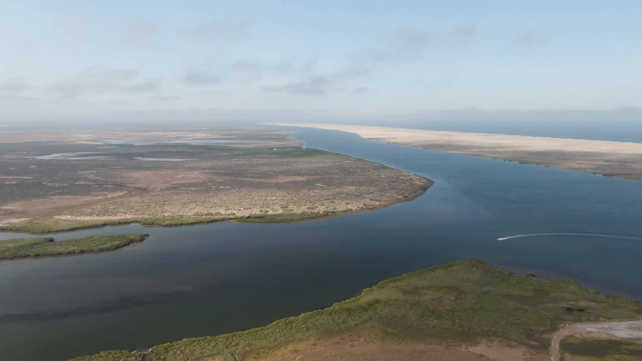 Aerial frame over Aldofo Lopez Mateos showing the waterway curving past low brown-green terrain toward a bright sand beach and ocean horizon, with no visible people.
