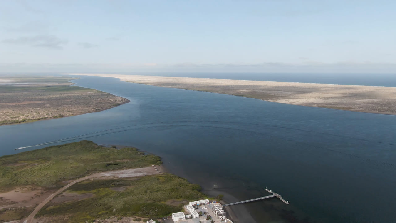 Aerial frame from Aldofo Lopez Mateos, Baja California Sur, showing blue lagoon water, green foreground shore, a dock with a small boat nearby, sandy land across the channel, and no visible people.