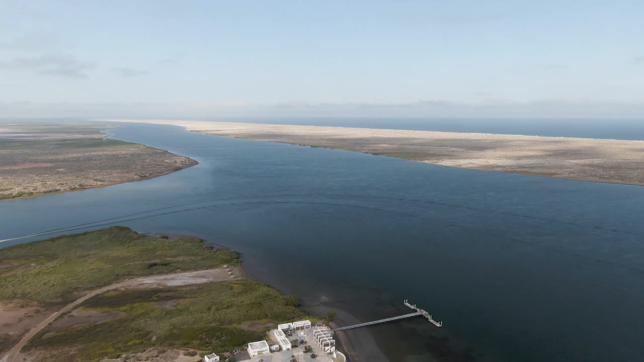 Second aerial frame over Aldofo Lopez Mateos water with the dock and boat lower in frame, dark ripples across the lagoon, sandy flats in the distance, and no visible people.
