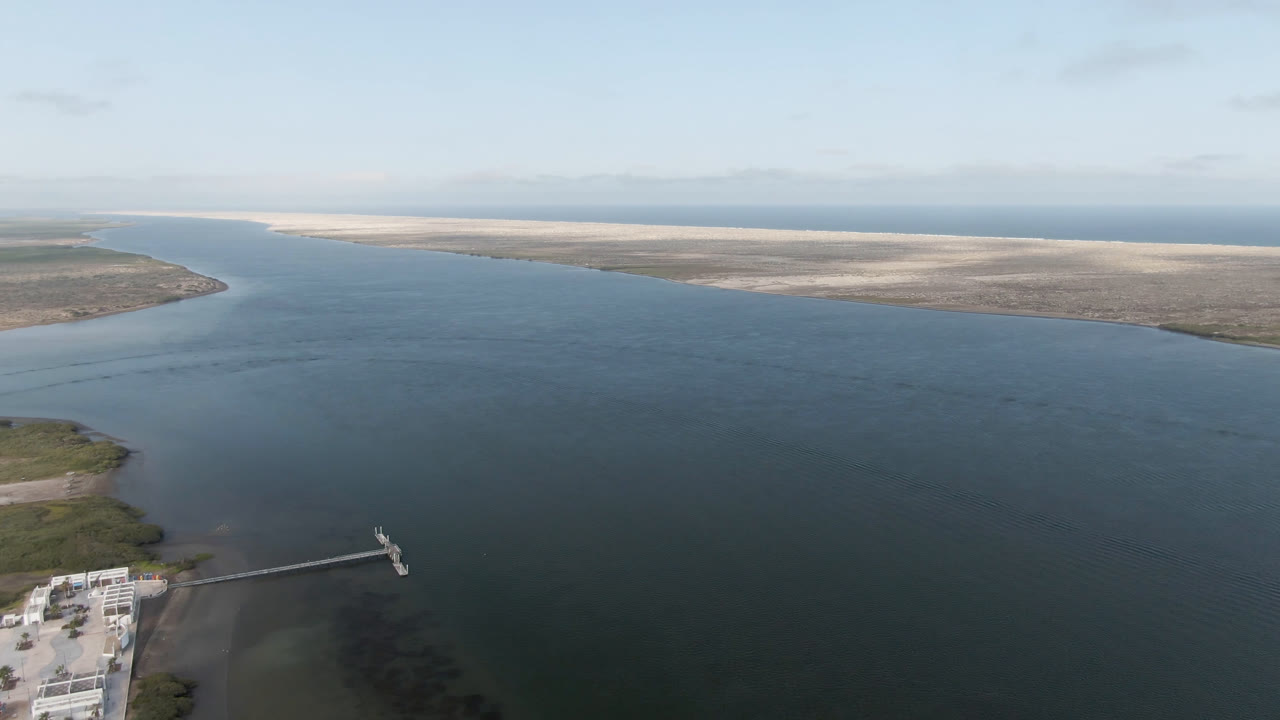 Wide aerial frame over Aldofo Lopez Mateos, Baja California Sur, with blue-green water in front, pale sandbars on the horizon, a dock edge at lower left, and no visible people.