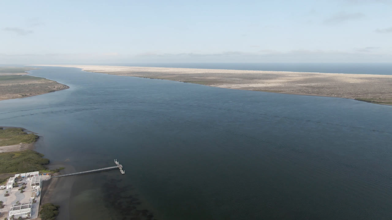 Aerial frame from Aldofo Lopez Mateos, Baja California Sur, showing the long pier more clearly at lower left, broad still water across the center, pale sandbars beyond, and no visible people.