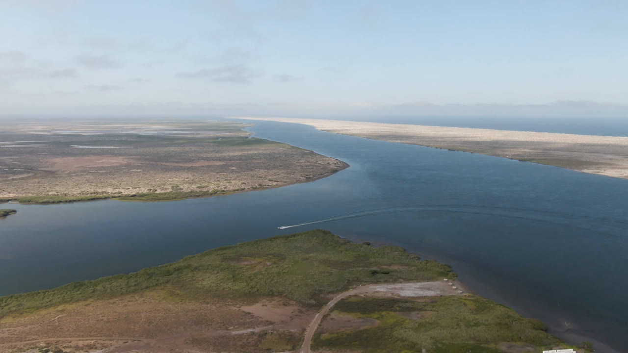 Wide aerial view of blue water, scrubland, sandbars, and a small boat wake.