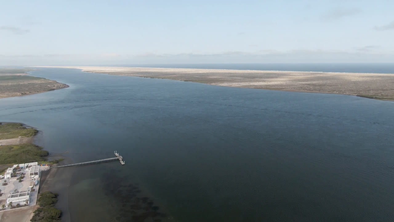 Aerial frame over Aldofo Lopez Mateos, Baja California Sur, with blue-green water dominating the view, a dock and pale buildings at lower left, distant sandy landforms on the horizon, and no visible people.
