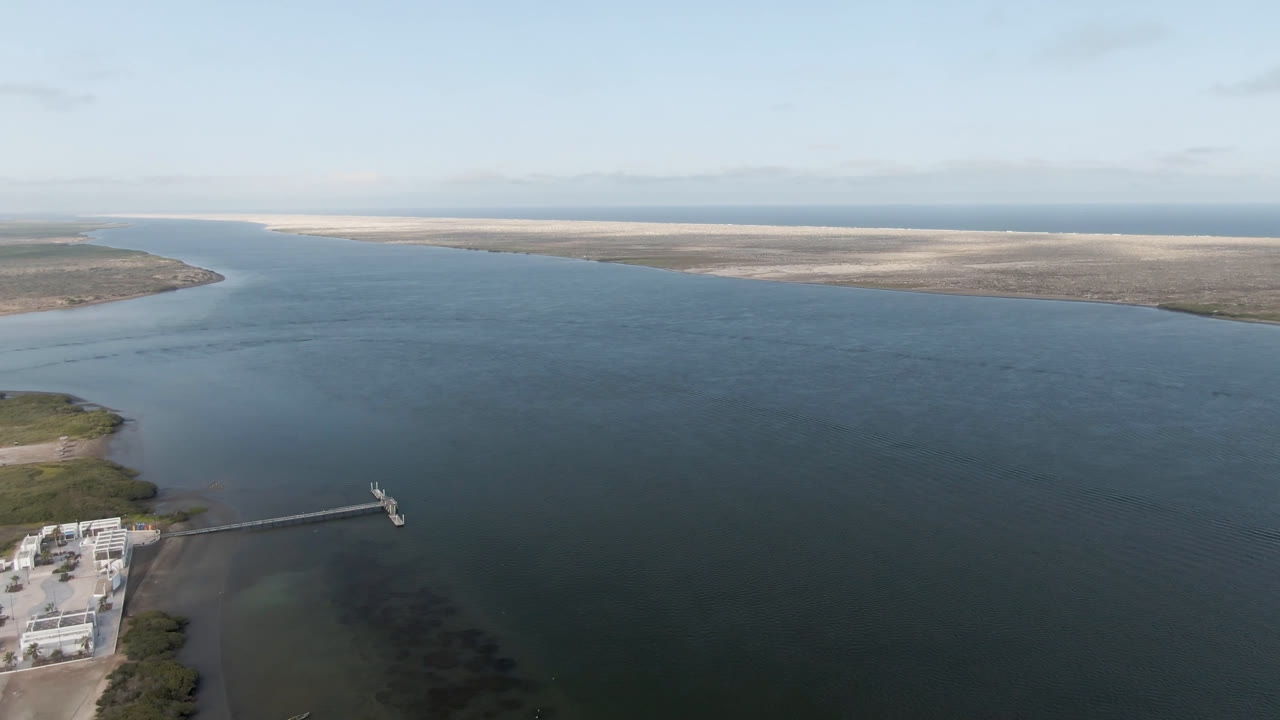 Aerial view in Aldofo Lopez Mateos showing the dock extending farther into the blue-green water from the lower-left shoreline, with low sandy land and a pale sky beyond, and no visible people.